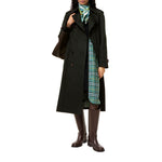 A model wearing a long dark green trench coat over a green and blue plaid dress, accessorized with a green and white silk scarf and dark brown knee-high boots, and carrying a dark brown shoulder bag, set against a plain white background.