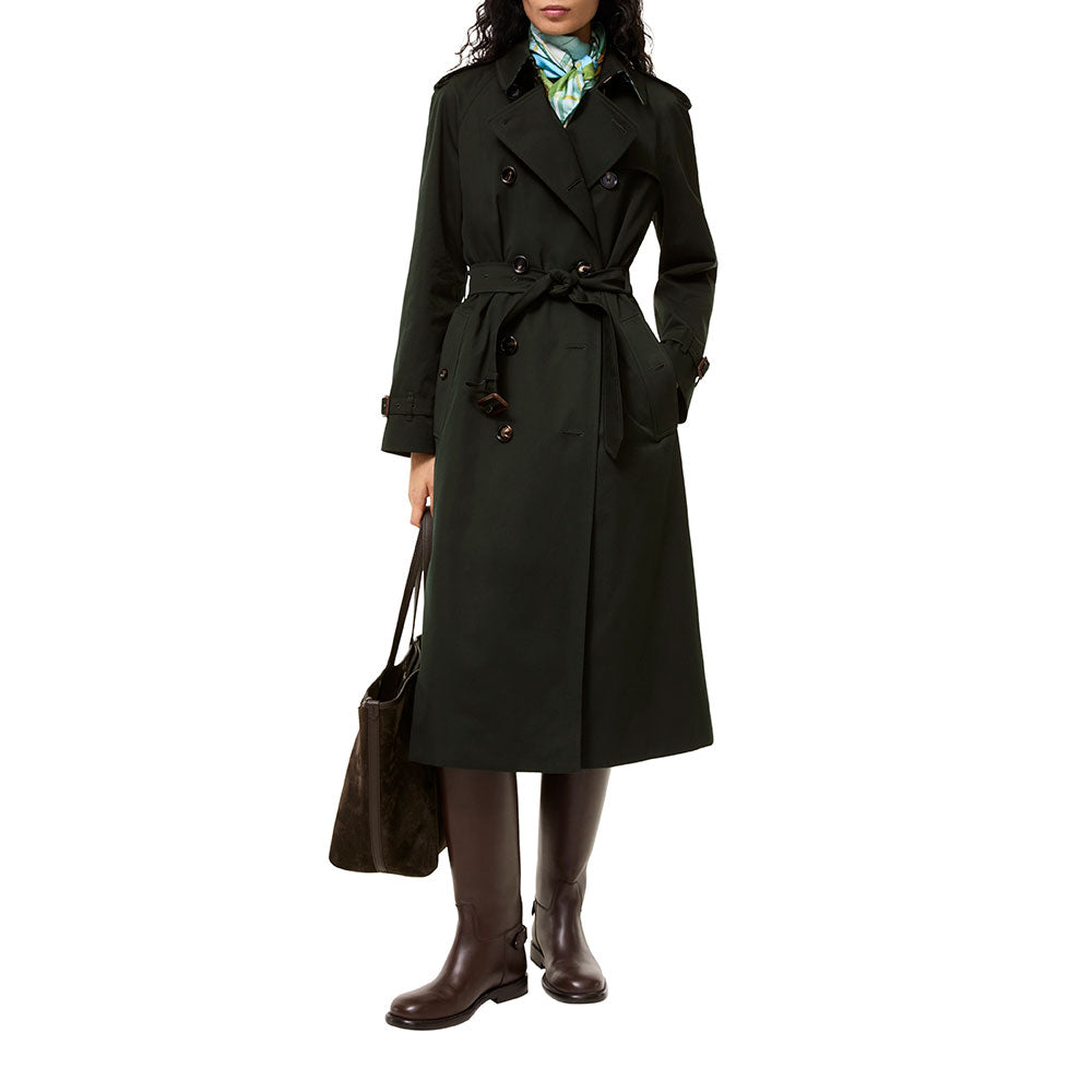 A woman modeling a long dark green trench coat with a matching waist tie, styled with a patterned silk scarf, brown leather knee-high boots, and a brown tote bag against a plain white background.
