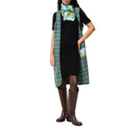 A model wearing a black dress with a green and white plaid vest draped over the shoulders, a matching patterned scarf, and brown knee-high boots, set against a plain white background.