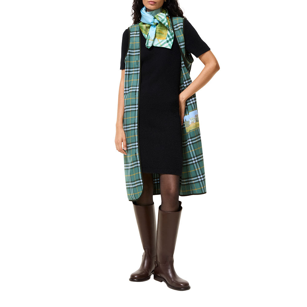 A model wearing a black dress with a green and white plaid vest draped over the shoulders, a matching patterned scarf, and brown knee-high boots, set against a plain white background.