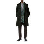 A model wearing a dark green mid-length overcoat over a gray sweater, a green and black plaid scarf, dark brown trousers, and black leather shoes, shot against a solid white background.