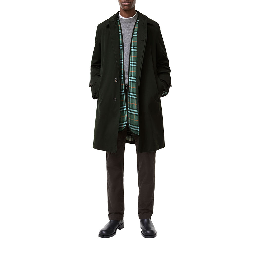 A model wearing a dark green mid-length overcoat over a gray sweater, a green and black plaid scarf, dark brown trousers, and black leather shoes, shot against a solid white background.