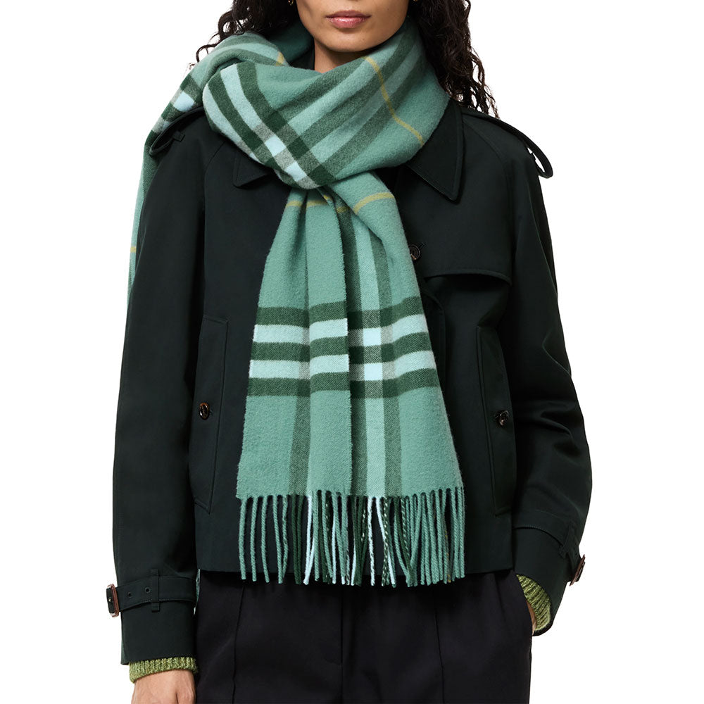 A model wearing a classic green plaid scarf with white and yellow stripes, styled over a dark green trench coat against a plain white background.