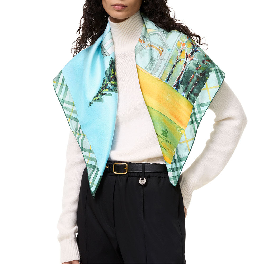 A model wearing a light blue and green plaid silk scarf with a printed landscape panel and a yellow-green section, styled over a white turtleneck sweater and black trousers.