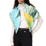 A model wearing a light blue and green plaid silk scarf with a printed landscape panel and a yellow-green section, styled over a white turtleneck sweater and black trousers.