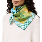 A close-up of a model wearing a Burberry silk scarf tied around her neck. The scarf features a green and yellow gradient with a plaid pattern and the brand name 'Burberrys' printed on it.