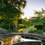 A serene landscaped garden at sunset with a stone path leading through neatly trimmed hedges and trees toward a house in the distance, with a curved reflecting pool in the foreground.