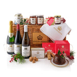 A Highgrove Christmas hamper gift set arranged in a wicker basket on a white studio background. The set includes a bottle of champagne, a bottle of red wine, a bottle of white wine, jars of preserves, a white waffle towel, boxed and tinned Christmas pudding, cookies, and holly decorations.