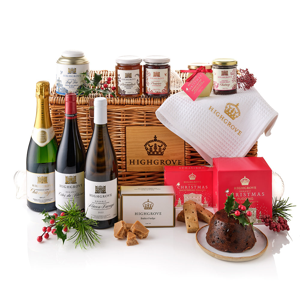 A Highgrove Christmas hamper gift set arranged in a wicker basket on a white studio background. The set includes a bottle of champagne, a bottle of red wine, a bottle of white wine, jars of preserves, a white waffle towel, boxed and tinned Christmas pudding, cookies, and holly decorations.
