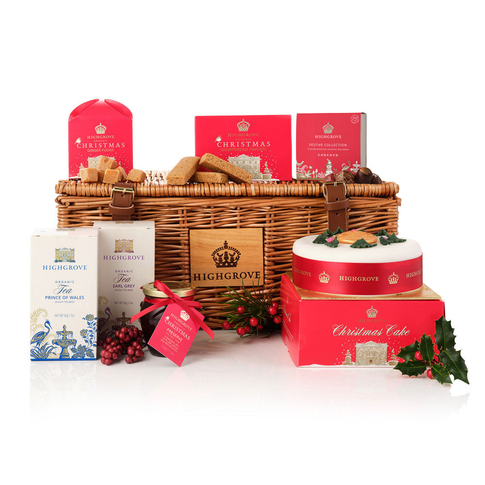A Highgrove Christmas hamper gift set on a white background, featuring a wicker basket with a branded wooden front panel, red Christmas boxes and treats, tea boxes, a decorated Christmas cake, and holly accents.