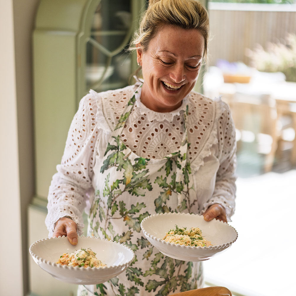 Kelly Bailey smiling while holding two white scalloped bowls filled with food, wearing a white eyelet blouse and a green botanical apron in a bright kitchen setting.
