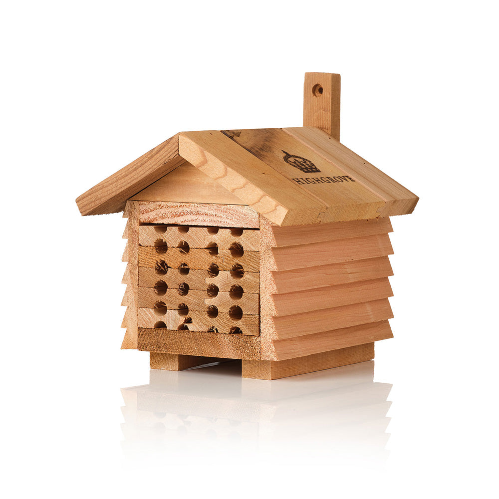 A small wooden bee hotel with a slatted front and a chimney, shown on a clean white background with a soft reflection.