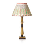 A table lamp with a cream-colored fabric shade decorated with a colorful hand-painted leaf pattern in green, red, and blue, supported by a turned wooden base in a warm gold finish with dark brown accents, set against a plain white background.