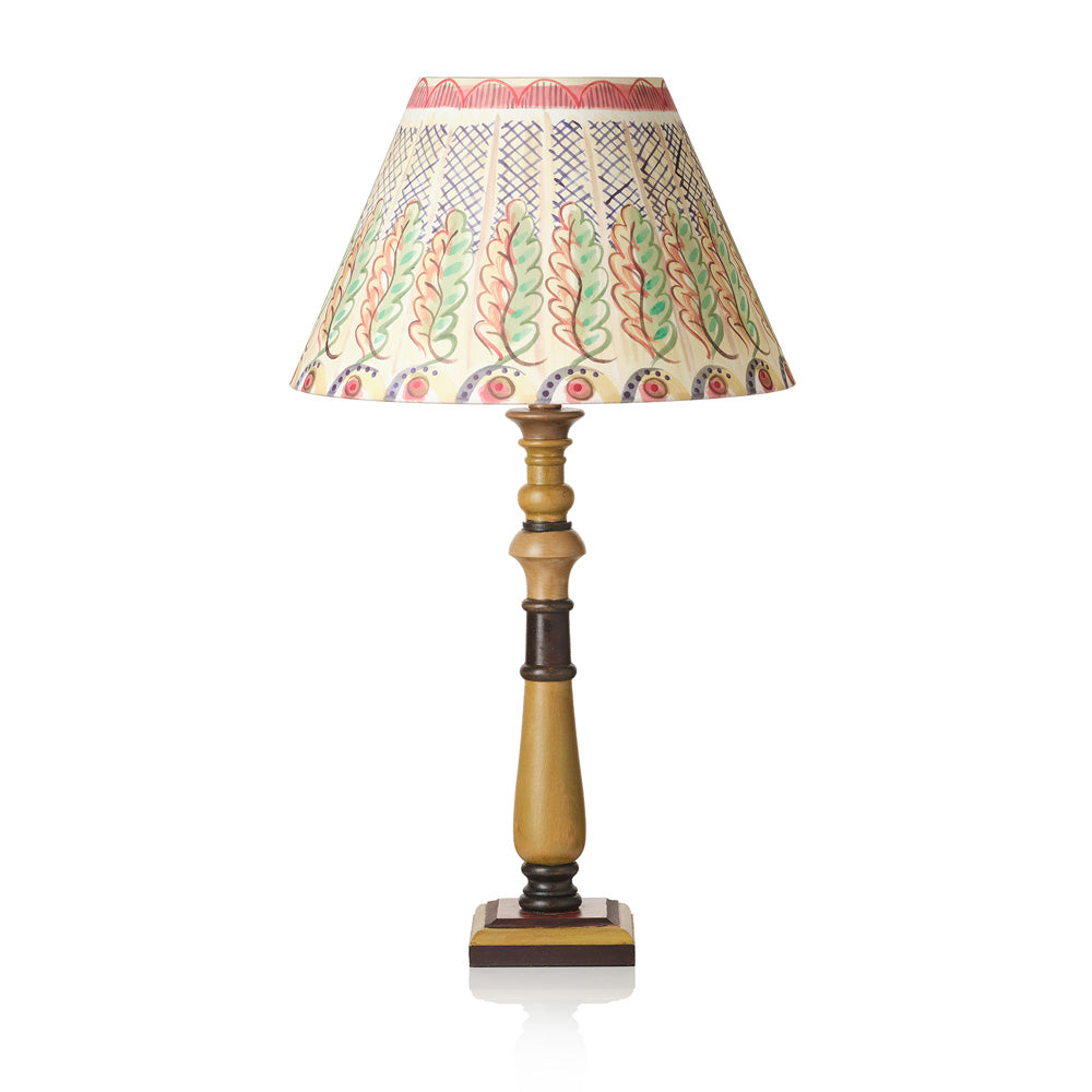 A table lamp with a cream-colored fabric shade decorated with a colorful hand-painted leaf pattern in green, red, and blue, supported by a turned wooden base in a warm gold finish with dark brown accents, set against a plain white background.