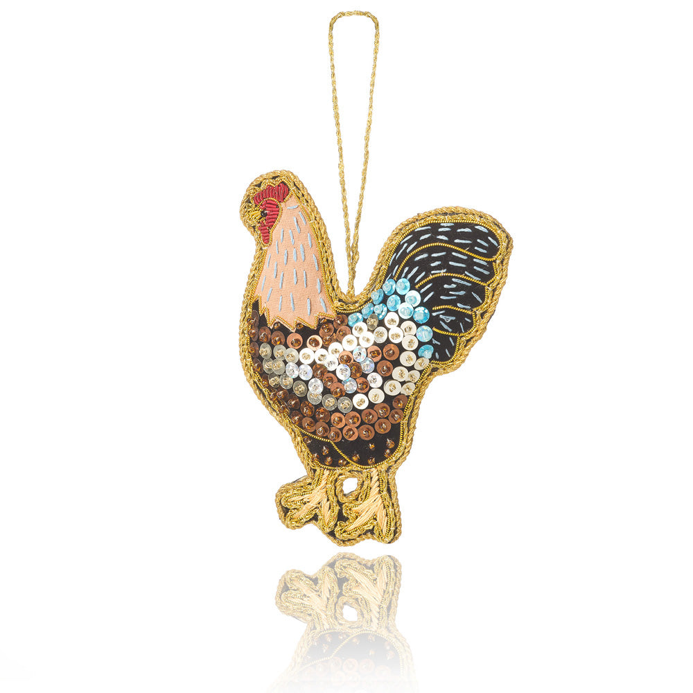 A decorative hanging ornament shaped like a rooster, featuring a gold embroidered outline, a light peach neck, a red comb, and a body covered with sequins in black, brown, white, and light blue. A gold hanging loop is attached at the top, shown on a white background with a soft reflection below.
