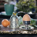 A decorative silver egg cup and matching silver cloche with a honeycomb pattern and gold butterfly accent, styled with eggs on a gold-trimmed tray in a softly blurred kitchen setting.