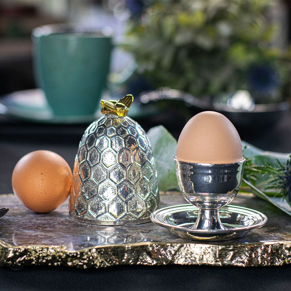 A decorative silver egg cup and matching silver cloche with a honeycomb pattern and gold butterfly accent, styled with eggs on a gold-trimmed tray in a softly blurred kitchen setting.
