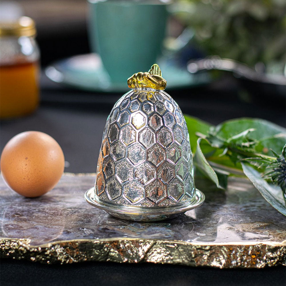 A decorative silver cloche with a honeycomb pattern and a gold frog topper, styled on a gold-rimmed tray with a brown egg and green leaves in a kitchen setting.