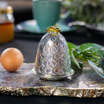 A decorative silver cloche with a honeycomb pattern and a gold frog topper, styled on a gold-rimmed tray with a brown egg and green leaves in a kitchen setting.