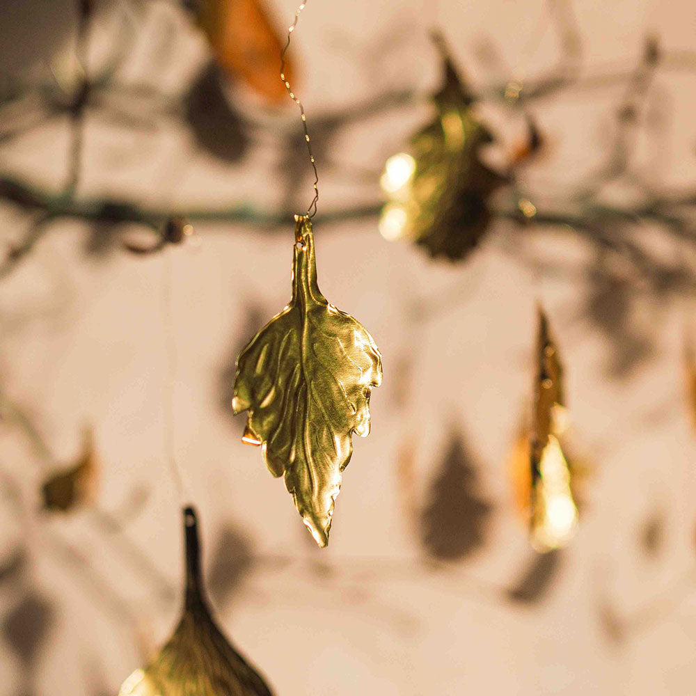 A close-up of a metallic gold leaf-shaped hanging ornament suspended from a thin wire, with other similar ornaments blurred in the background against a warm beige wall.