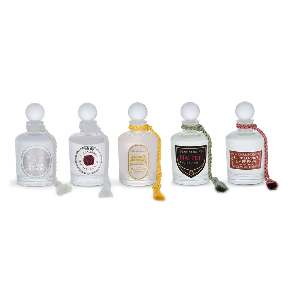 Five clear glass perfume bottles with white stoppers arranged in a row on a white background. Each bottle has a round or shield-shaped label with different colors and text, and some bottles have decorative cords with tassel ends.