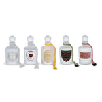 Five clear glass perfume bottles with white stoppers arranged in a row on a white background. Each bottle has a round or shield-shaped label with different colors and text, and some bottles have decorative cords with tassel ends.