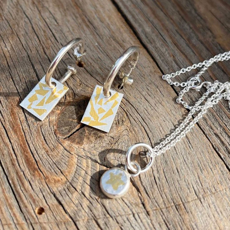 Silver jewellery set featuring a pair of hoop earrings with rectangular white and gold geometric charms, and a matching silver chain necklace with a round white and gold pendant, displayed on a rustic wooden surface.